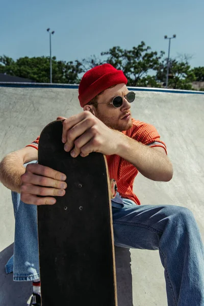 young and trendy man in sunglasses sitting with skateboard and looking away