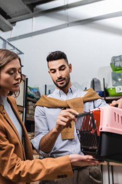 Muslim man holding animal cage near blurred girlfriend in pet shop