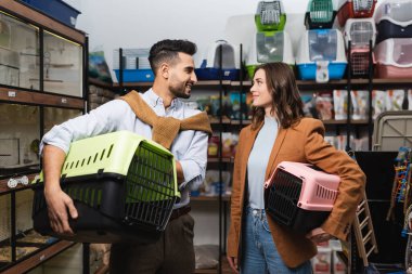 Cheerful multiethnic couple holding animal cages in pet shop 