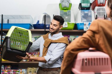 Smiling muslim man holding animal cage near blurred girlfriend in pet shop
