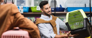 Cheerful muslim man looking at animal cage near blurred woman in pet shop, banner 