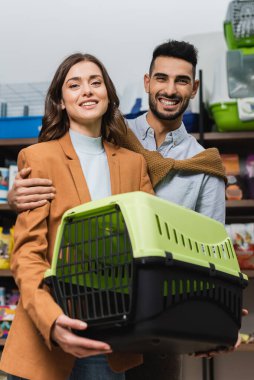 Smiling multiethnic couple holding animal cage and looking at camera in pet shop