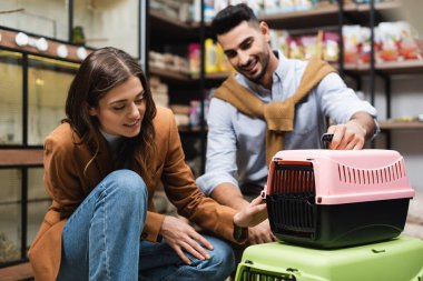 Smiling woman choosing animal cage near blurred muslim boyfriend in pet shop 