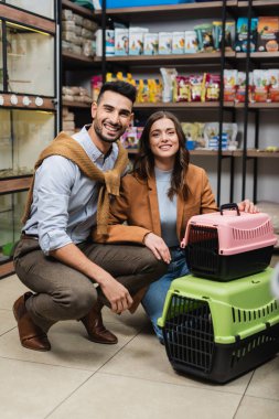 Cheerful interracial couple looking at camera near cages in pet shop 