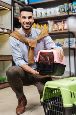 Cheerful arabian man holding pet cage and looking at camera in pet shop 