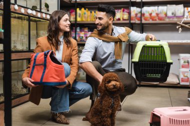 Smiling multiethnic couple holding bag and cage near poodle in pet shop