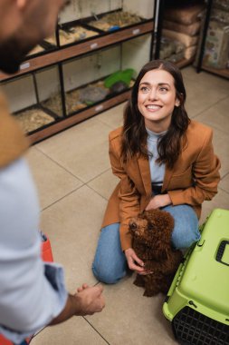 Smiling woman petting poodle near cage and blurred boyfriend in pet shop