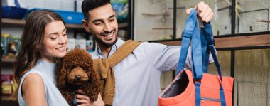 Muslim man holding animal dog near girlfriend with poodle in store, banner  clipart