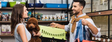 Side view of cheerful interracial couple holding poodle and animal cage in store, banner 