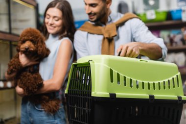 Blurred multiethnic couple with poodle holding animal cage in shop 