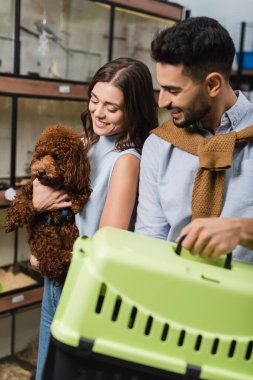 Smiling woman holding poodle near arabian boyfriend with animal cage in pet shop 