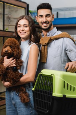 Cheerful multiethnic couple holding poodle and animal cage in pet shop 