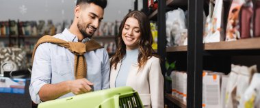 Smiling muslim man holding animal cage near girlfriend and showcase in animal shop, banner 