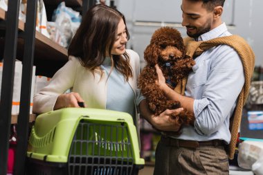 Smiling interracial couple looking at poodle and holding animal cage in pet shop 