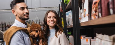 Multiethnic couple with poodle looking at packages in pet shop, banner 