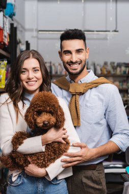 Cheerful woman holding poodle near muslim boyfriend in pet shop 