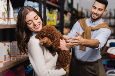 Smiling woman holding poodle near blurred arabian boyfriend and packages in pet shop 