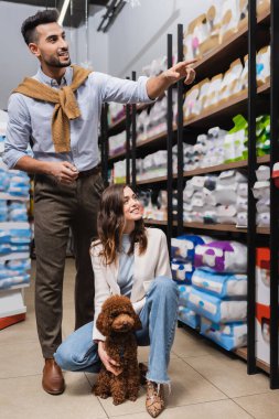 Smiling muslim man pointing at packages near girlfriend with poodle in pet shop 