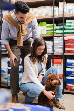 Smiling muslim man looking at girlfriend with poodle in pet shop 