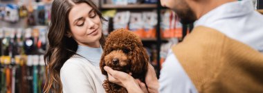 Woman holding poodle near blurred man in pet shop, banner 