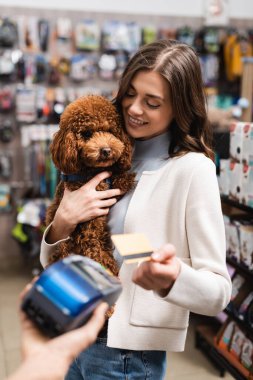 Smiling woman looking at poodle and paying with credit card in pet shop 