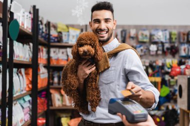 Positive arabian man holding poodle and looking at camera while paying with credit card in pet shop  clipart