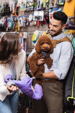 Cheerful interracial couple with poodle choosing animal jacket in pet shop 