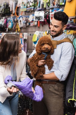 Cheerful interracial couple with poodle choosing animal jacket in pet shop  clipart