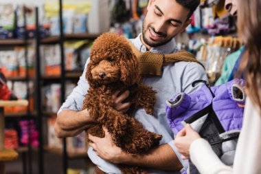 Blurred woman holding animal jacket near arabian boyfriend with brown poodle in pet shop 
