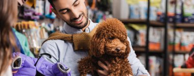 Smiling arabian man looking at poodle near girlfriend with animal jacket in pet shop, banner 