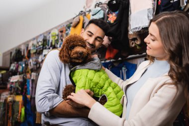 Smiling interracial couple wearing animal jacket on poodle in animal shop 