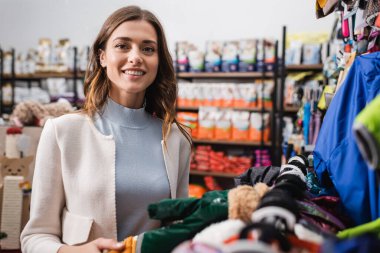 Cheerful woman looking at camera near animal clothes in pet shop clipart