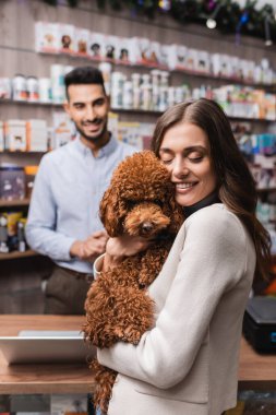 Smiling woman hugging poodle near blurred muslim seller in pet shop 