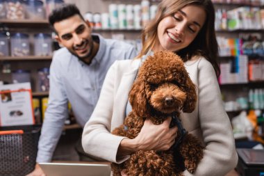 Smiling woman holding poodle near blurred arabian salesman in pet shop