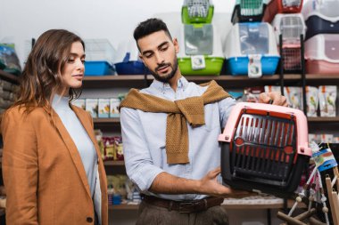 Arabian man holding animal cage near brunette girlfriend in pet shop