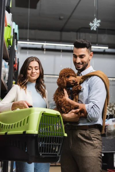 Cheerful arabian man holding poodle near girlfriend with animal cage in pet shop 