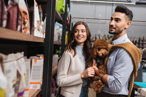 Interracial couple with poodle looking at packages on showcase of pet shop 