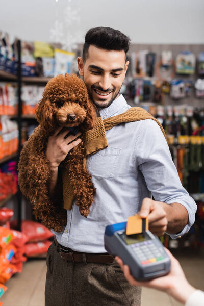 Cheerful muslim man holding poodle and paying with credit card near seller in pet shop 