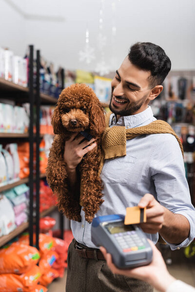 Smiling arabian man looking at poodle while paying with credit card in pet shop