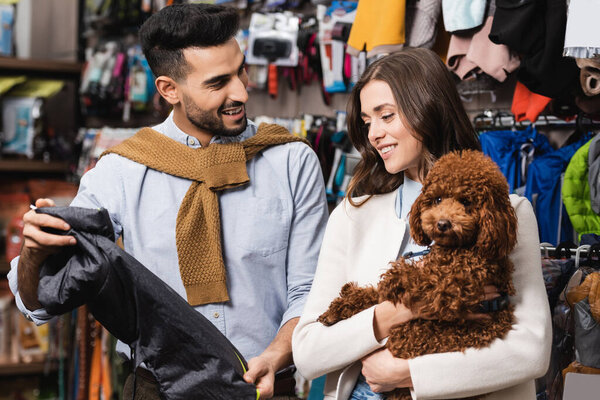 Арабский мужчина держит пиджак рядом с подружкой с пуделем в зоомагазине 