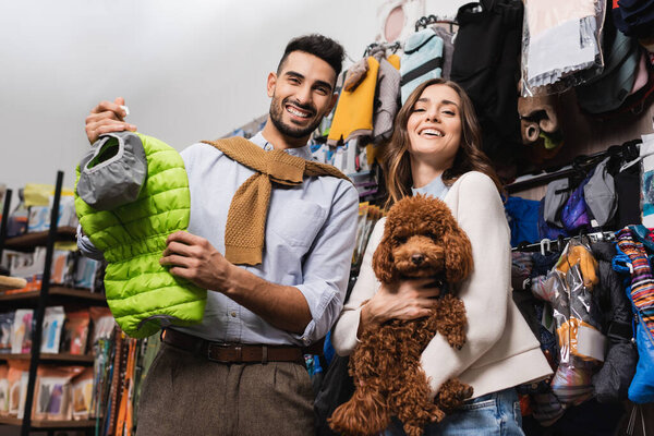 Низкий угол обзора веселой межрасовой пары, держащей пуделя и пиджак в зоомагазине 
