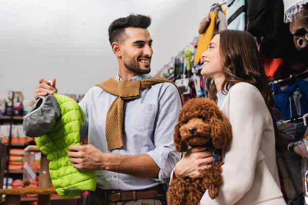 Cheerful multiethnic couple holding poodle and animal jacket in pet shop 