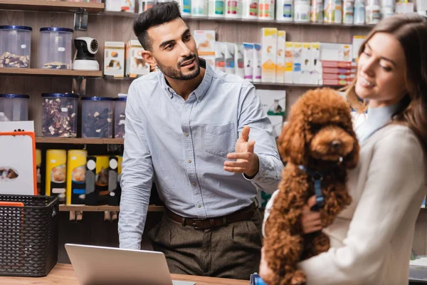 Muslim Salesman Pointing Blurred Woman Poodle Pet Shop — ストック写真