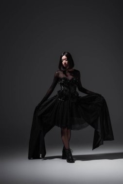 full length of asian woman in black gothic dress and stylish boots posing on dark grey