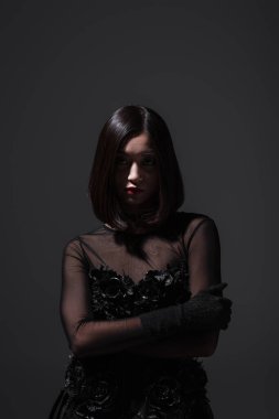 brunette asian woman in black gothic dress and gloves posing with crossed arms isolated on dark grey