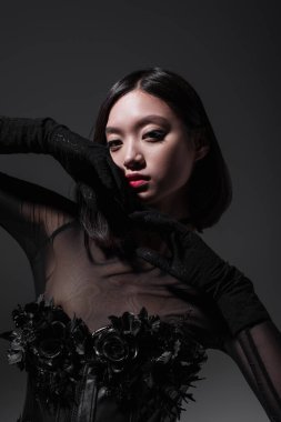 brunette asian woman in black gothic outfit and gloves posing isolated on dark grey