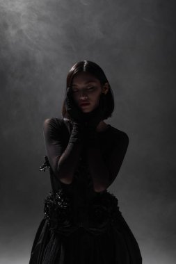 brunette asian model with closed eyes posing in black gothic outfit on dark grey with smoke