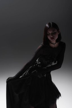 young asian woman in black gothic dress holding lace skirt while posing on grey
