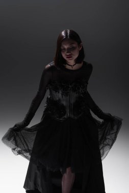 asian model in black gothic dress holding lace skirt while posing on grey