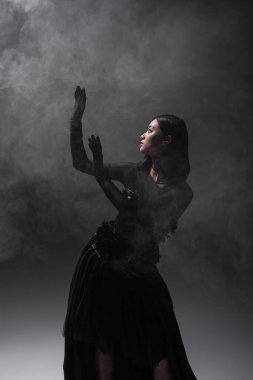 asian woman in gloves and gothic dress gesturing on grey with smoke
